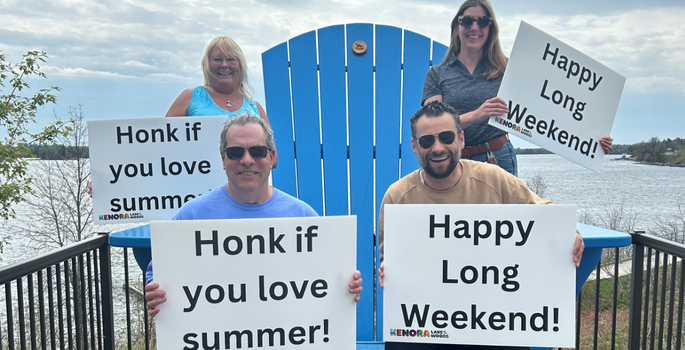 people sitting on an oversized blue Adirondack chair holding signs to welcome visitors